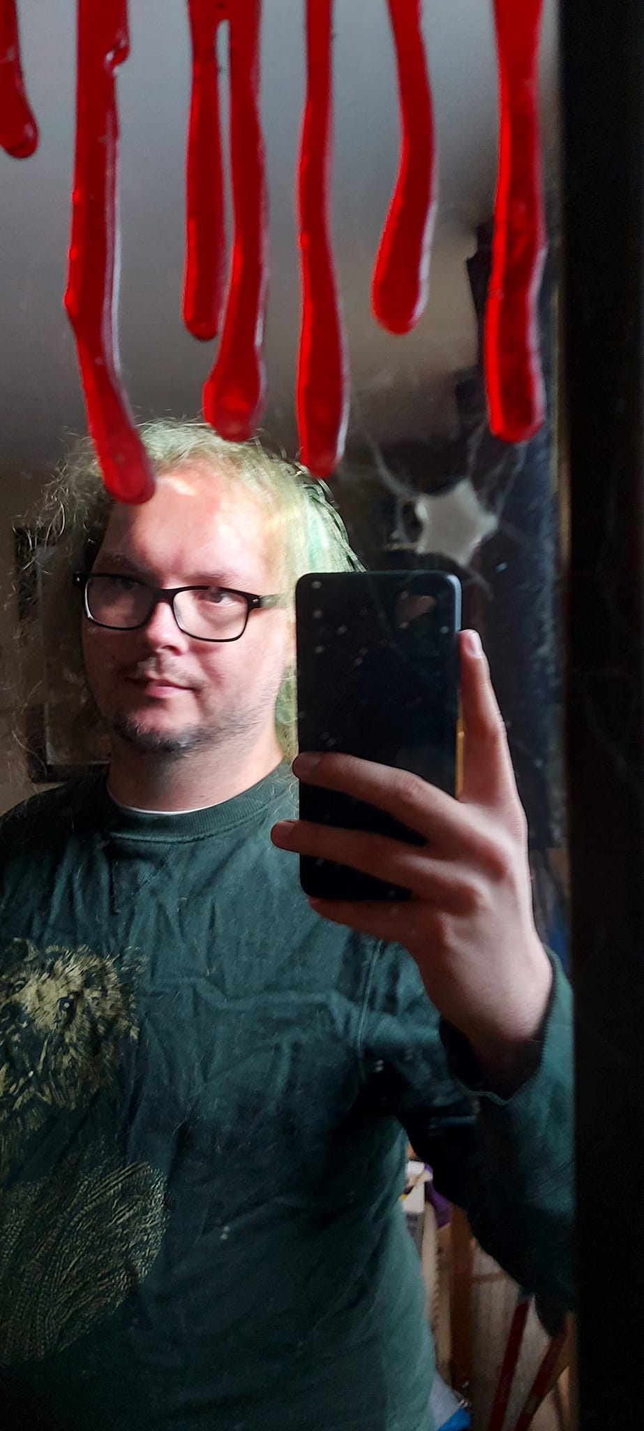 A man with long green hair wearing a sweater with a bear wearing a sweater on it stands in front of a mirror with a a window sticker that looks like dripping blood on it